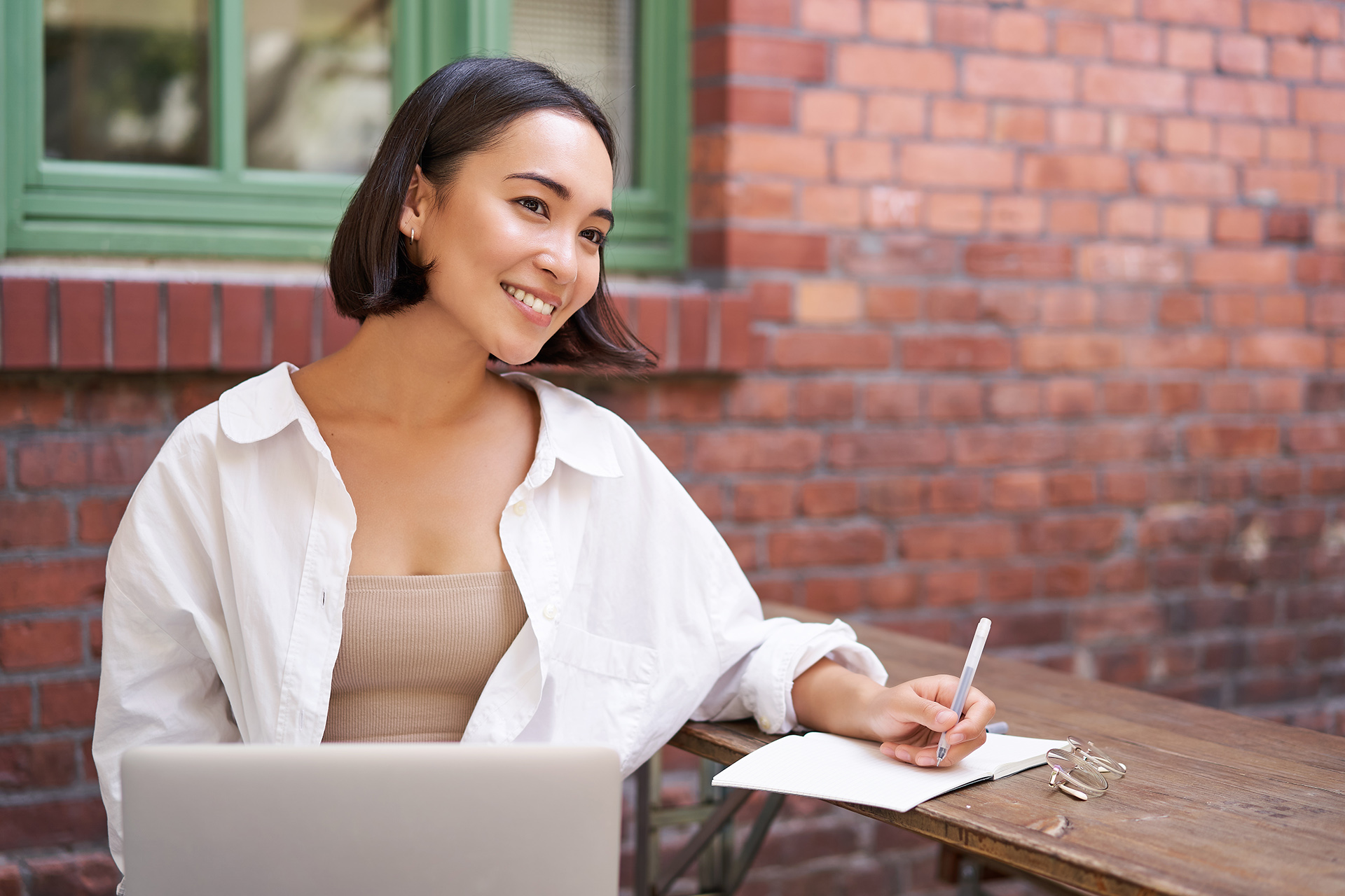 Beautiful Young Female Model Sitting With Laptop In Outdoor Cafe, Taking Notes, Writing In Her Diary Or Working On Project, Doing Homework